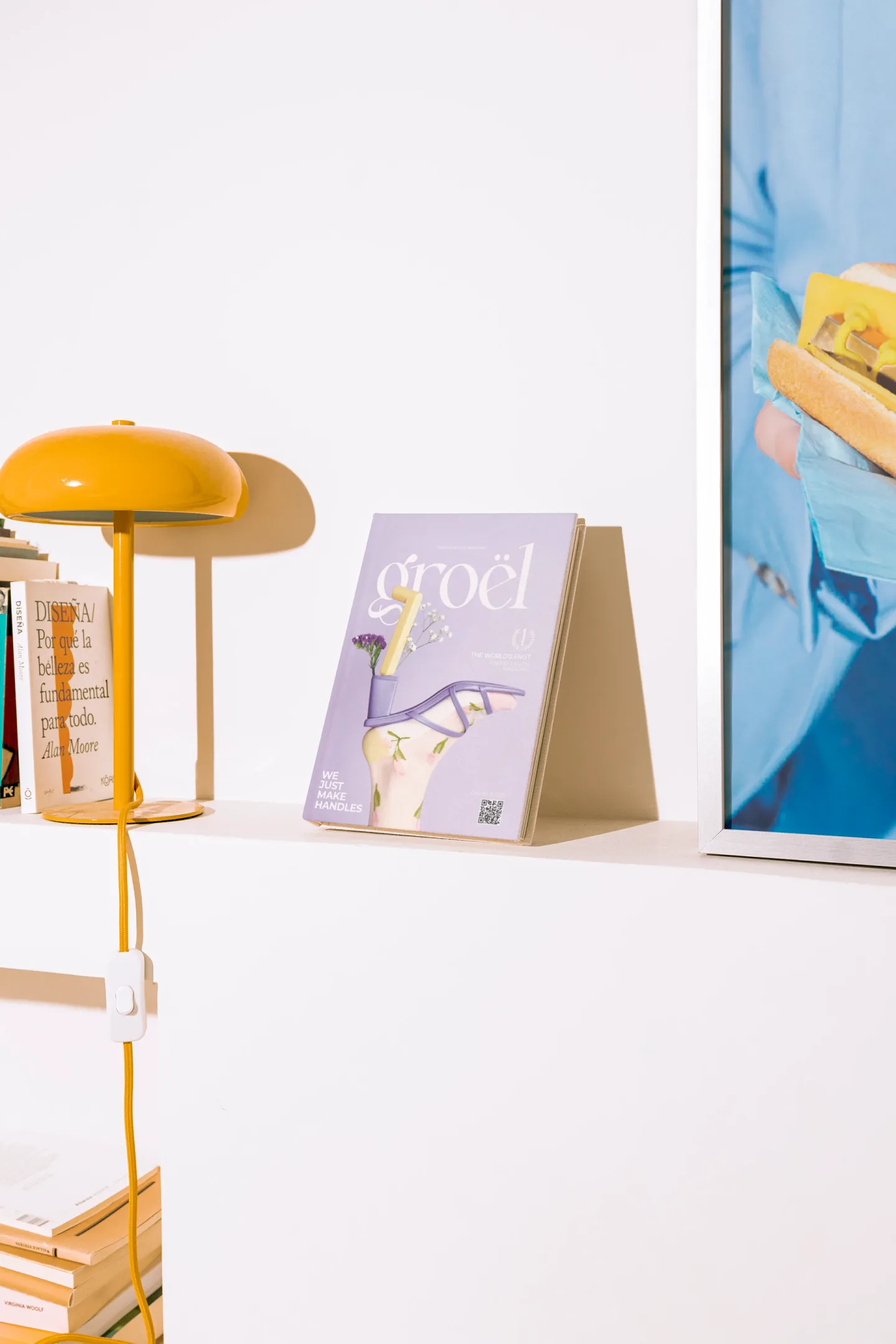 The Magazine en una estantería: portada lila con una sandalia de tacón hecha con una manilla, junto a libros y una lámpara mostaza